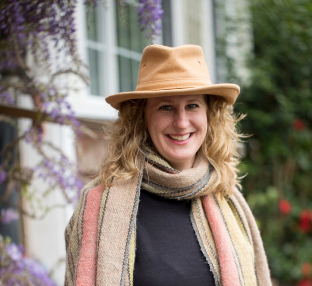 A smiling white woman wears a brown-cotton hat and pastel-striped scarf in front of hanging purple wisteria flowers
