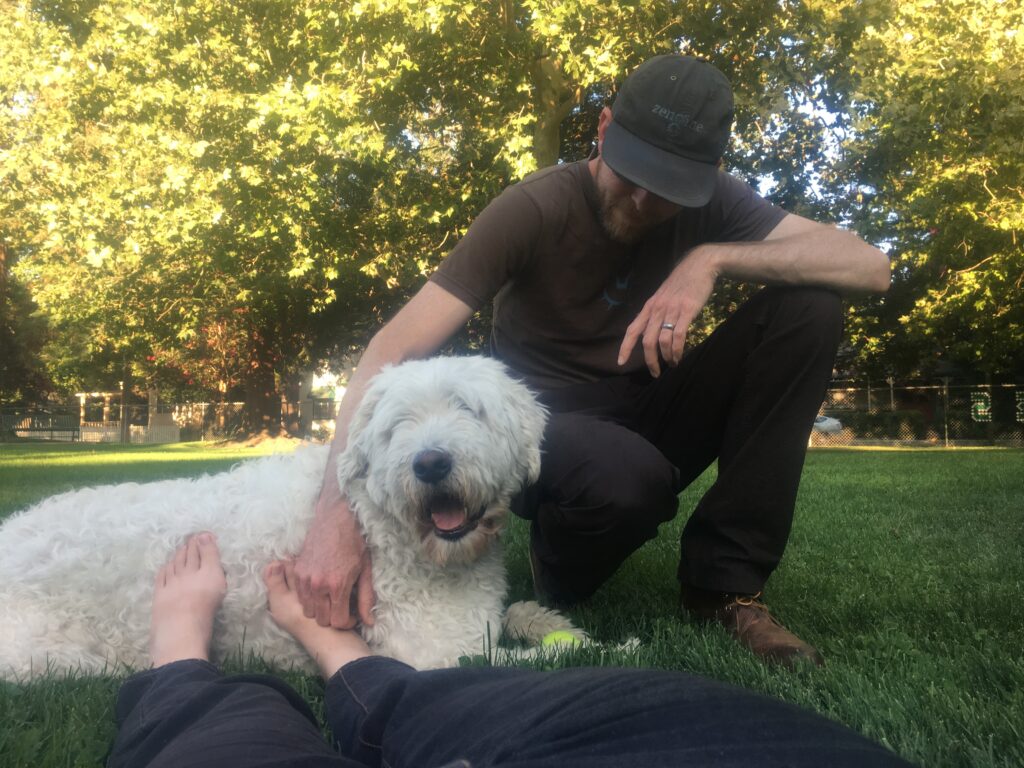 A white man in a baseball cap kneels with his right arm on a big white dog who is laying on green grass